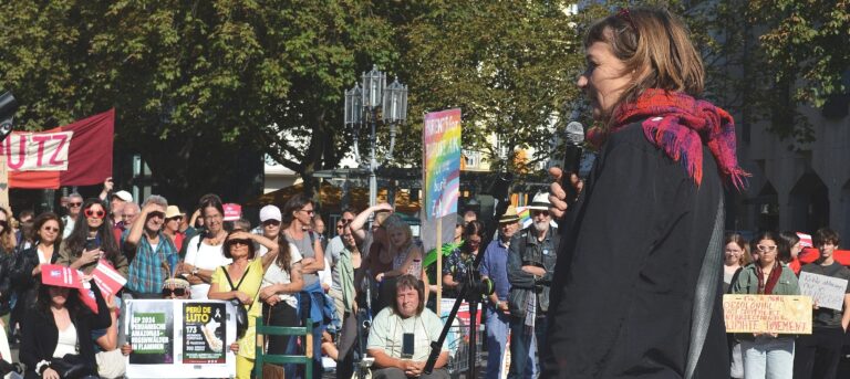 Gesa Maschkowski von hinten auf der Bühne vor Demonstrant_innen mit Schildern