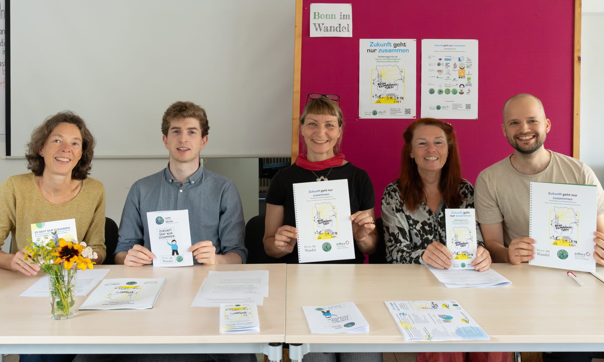 5Personen sitzen vor einer Pinwand hinter einem Tisch und halten Flyer und Ausdrucke des Zukunftsprogramms in die Kamera. Foto Ingrid Blessing