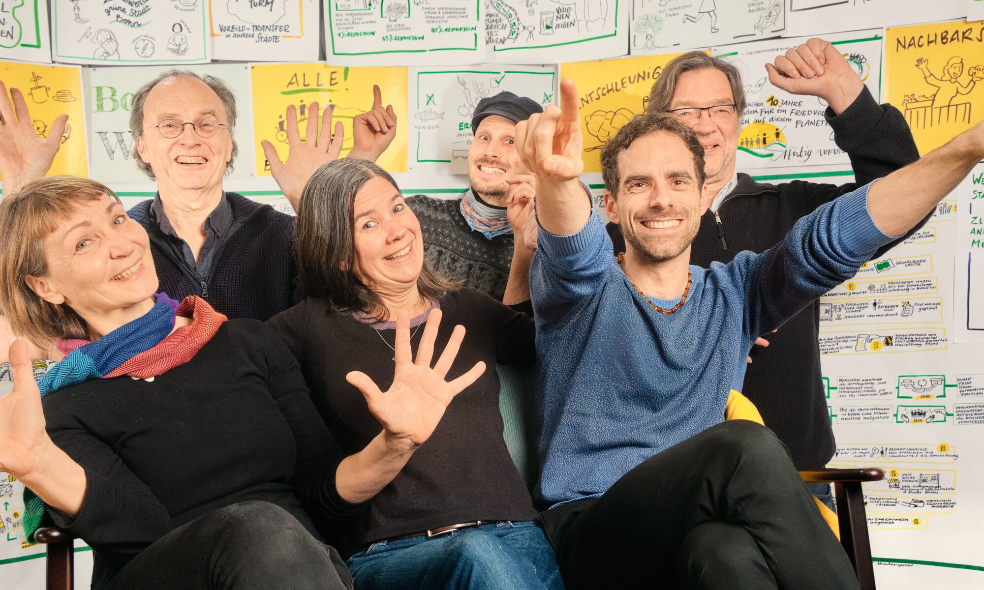 Das Foto zeigt sechs Personen, die sich sichtbar freuen. Sie sitzen vor einer Wand voller Illustration für eine schöne gerechte und soziale Zukunft, Foto Nele Lilly Schnüll