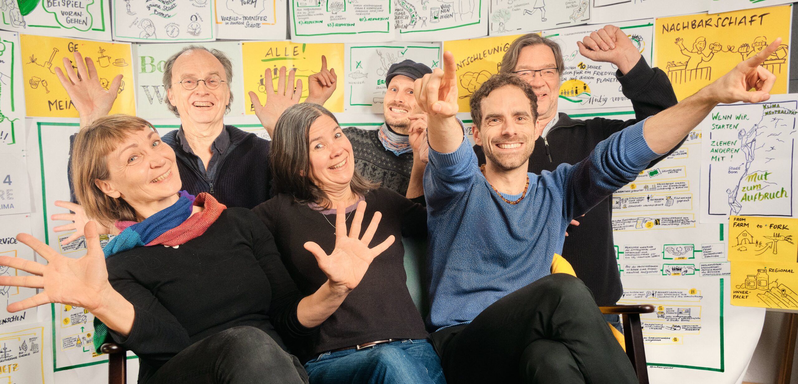 Das Foto zeigt sechs Personen, die sich sichtbar freuen. Sie sitzen vor einer Wand voller Illustration für eine schöne gerechte und soziale Zukunft