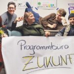 Die Gründer_innen des Programmbüros Zukunft stralen in die Kamera und halten ein großes Plakat hoch auf dem Programmbüro Zukunft steht