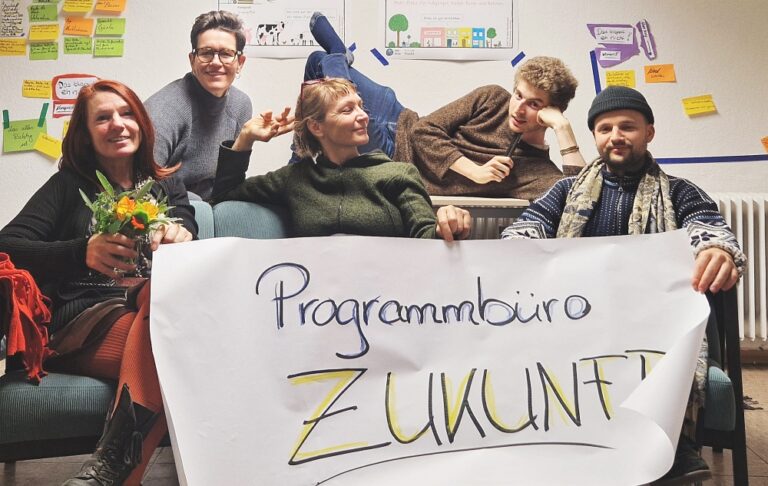 Die Gründer_innen des Programmbüros Zukunft stralen in die Kamera und halten ein großes Plakat hoch auf dem Programmbüro Zukunft steht