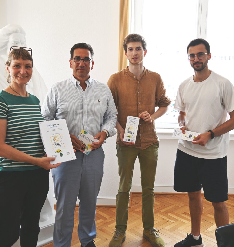 Gesa Maschkowski, Guido Déus, Raphael Räpple und Max Frenken stehen im Büro von Guido Deus und halten Flyer vom Zukunftsprogramm in den Händen und lächeln freundlich in die Kamera