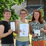 3 Personen in sommerlicher Kleidung stehen unter einem Baum und halten lächelnd das Zukunftsprogramm in die Kamera