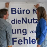Ein Mann undn eine Frau hängen ein großes Banner auf. Darauf steht: Büro für die Nutzung von Fehlern und Zufällen