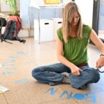 Eine Frau mit Blonden Haaren und grünen Shirt sitzt im Schneidersitz auf dem Boden des Pavillon und arbeitet mit blauem Klebe Tape