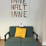 Über einem grünblauen Sofa mit gelbem Kissen hängt eine Leinwand auf der mit großen Buchstaben steht INNE HALT INNE