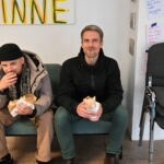 2 junge Männer sitzen auf dem Grünen Sofa. essen Fallafel und gucken in die Kamera