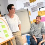 Workshopteilnehmende präsentiert ihre Ideen mit bunten Zetteln auf einer Leinwand, ein zweiter guckt zu