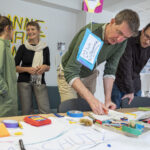 2 männliche Personen beugen sich über einen Materialtisch und füllen ein Zertifikat aus, 2 Personen im Hintergrund unterhalten sich