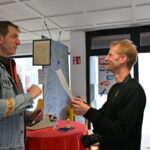 Ein großer Mann links in Jeans Jacker spricht zu ein einem etwas kleineren Mann der ein Dokument in der Hand hält