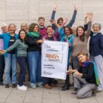 Menschen halten ein Flipchart-Plakat auf dem steht "Skills4Transformation". Sie stehen vor eine Wand und winken und lachen in die Kamera