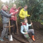 Fünf Personen mit Gartengeräten zeigen auf einen Komposthaufen