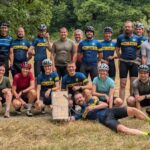 viele junge Menschen in Fahrradkluft strahlen in die Kamera. Sie stehen in und liegen in 2 Reihen im Sonnenschein